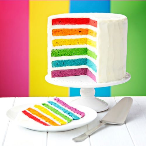 a rainbow layer cake with a slice cut out showing the rainbow coloured layers inside the white frosting